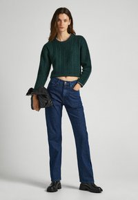 Maglione all'uncinetto verde con una vestibilità ampia e texture a coste, abbinato a pantaloni a vita alta a righe blu e stivaletti neri.