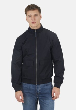 Młody mężczyzna zwrócony przodem, ubrany w granatową kurtkę na zamek z klapami i niebieskie dżinsy, na tle jednolitego jasnego tła.