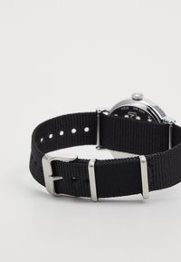 Cinturino in tessuto nero con fibbia e dettagli in metallo argentato. Presenta un quadrante rotondo con sfondo nero e dettagli argentati.