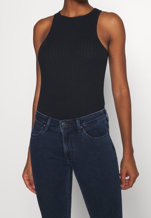 Vrouw draagt een mouwloos zwart geribbeld topje gecombineerd met donkerblauwe denim jeans, staand tegen een effen witte achtergrond.