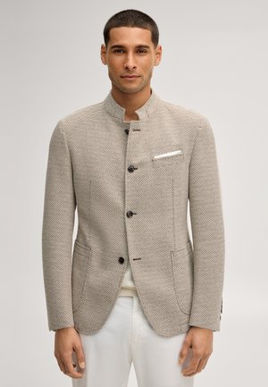 Mann trägt einen strukturierten beigen Blazer mit schwarzen Knöpfen und einem weißen Einstecktuch, kombiniert mit weißen Hosen vor einem schlichten Hintergrund.