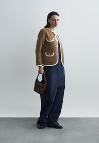 Brązowa wełniana kurtka z białymi lamówkami i kieszeniami; w zestawie z luźnymi granatowymi spodniami i bordową torebką. Czarne buty dopełniają strój.