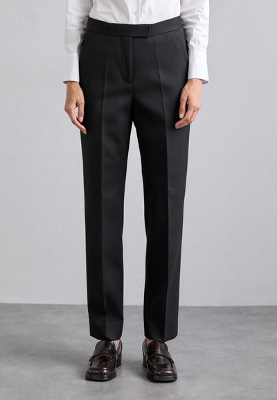 Pantalons noirs ajustés à l'avant plat, avec des poches latérales et une texture lisse, associés à des mocassins sombres et à une chemise blanche à boutons.