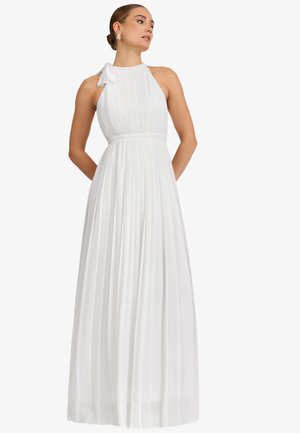 Frau trägt ein ärmelloses weißes plissiertes Maxikleid mit einer Schleifendetail an einer Schulter, steht mit den Händen hinter dem Rücken.