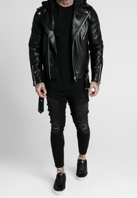 Veste de moto en cuir noir avec des fermetures éclair argentées, portée par-dessus un t-shirt noir. Jeans skinny noirs déchirés et baskets noires avec semelles blanches.