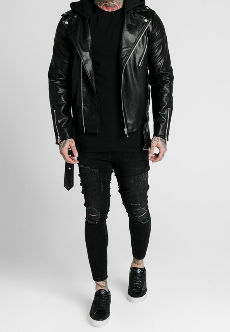 Veste de moto en cuir noir avec des fermetures éclair argentées, portée par-dessus un t-shirt noir. Jeans skinny noirs déchirés et baskets noires avec semelles blanches.