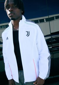 Hvid zip-up jakke med sorte striber og røde detaljer, Adidas-logo på brystet, Juventus-logo på siden, lavet af letvægtsmateriale.