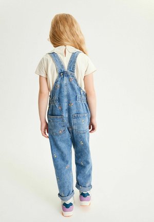 Peto vaquero con patrones florales bordados, tirantes ajustables y dos bolsillos en la parte trasera. Combinado con una camiseta de color claro y zapatillas deportivas coloridas.