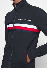 Námořnická zipová bunda s vysokým límcem, která má vodorovné pruhy v červené, bílé a námořnické barvě na hrudi a jemné detaily loga.