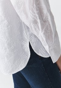 Blouse blanche brodée avec des motifs floraux, en tissu léger, manches bouffantes, ourlet arrondi, portée sur un jean en denim foncé.
