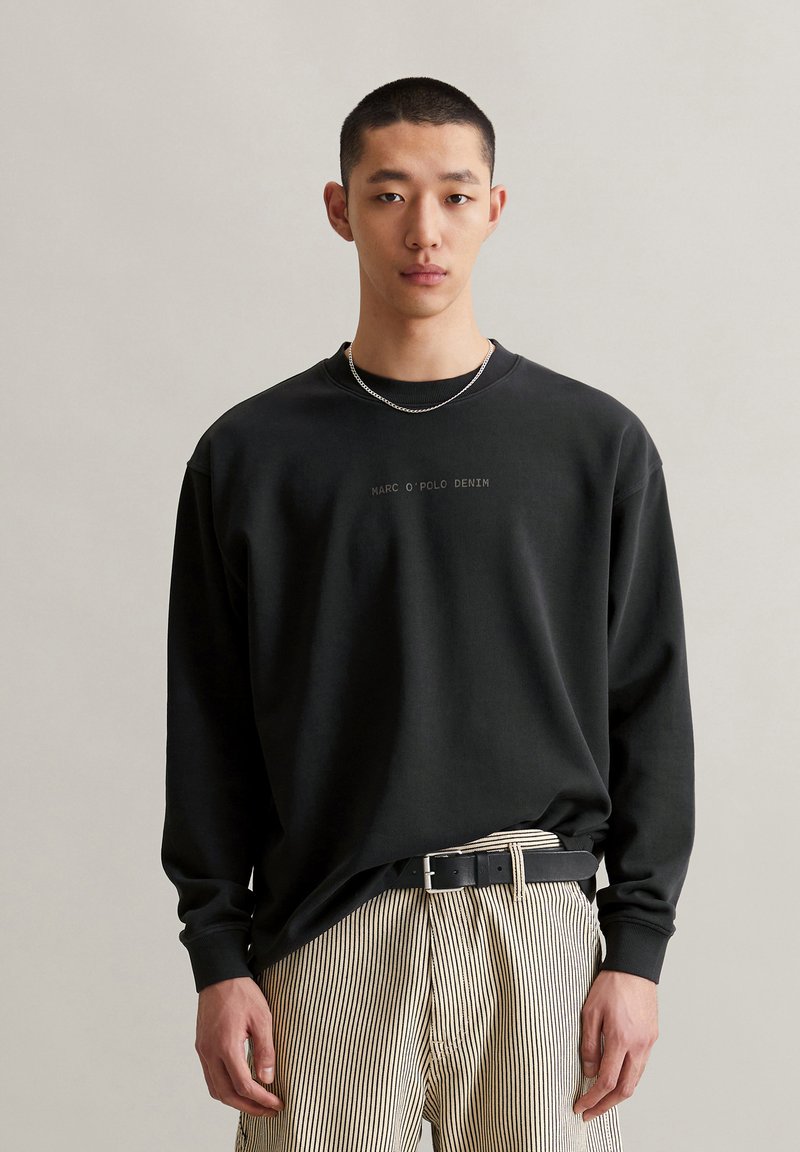 Schwarzer Pullover mit rundem Ausschnitt, verziert mit dezentem Branding-Text in der Mitte. Kombiniert mit beigen gestreiften Hosen und einem schwarzen Gürtel.