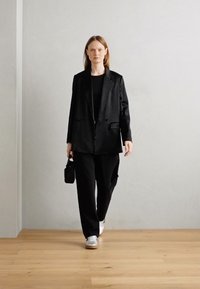 Blazer noir surdimensionné, top noir, pantalon large noir, baskets blanches. Le modèle tient un petit sac noir. Tissu lisse, design minimaliste.