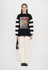 Black sweatshirt with striped sleeves, featuring a central printed graphic and stitched accents. Paired with cream wide-leg pants and black shoes.