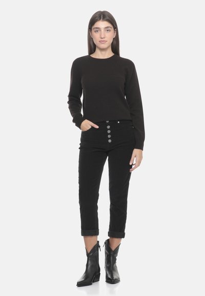 Maglione nero, pantaloni in velluto a coste neri con chiusura anteriore a cinque bottoni e stivaletti neri con tacco leggero. Texture liscia, design aderente.