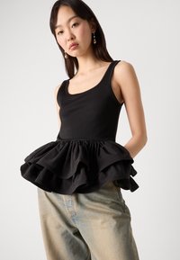 Čierny top na ramienka s volánovým peplum lemom, vyrobený z mäkkej tkaniny. Nosený s svetlomodrými džínsami. Model stojí proti šedému pozadiu.
