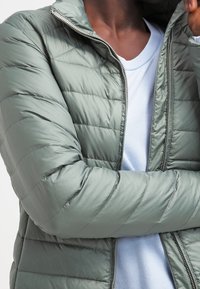 Lysegrøn, quilted pufferjakke med høj krave og lynlås, lagd over en lyseblå skjorte, som viser en glat, skinnende tekstur.