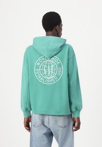 Hanorac verde cu glugă, având un design grafic alb pe spate care include un glob și textul „WORLDWIDE ESTABLISHED 1969.”