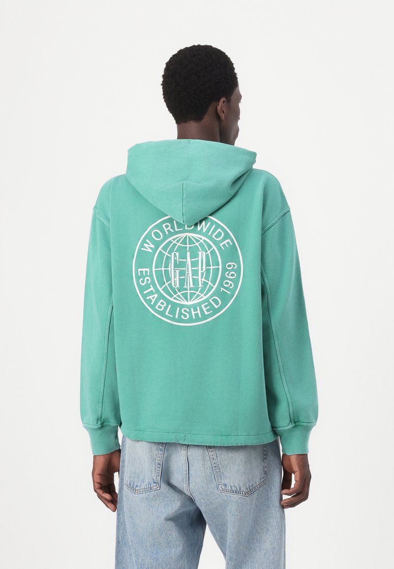 Hanorac verde cu glugă, având un design grafic alb pe spate care include un glob și textul „WORLDWIDE ESTABLISHED 1969.”
