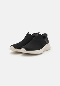Fekete slip-on sportcipők, texturált kötött felsőrésszel és kontrasztos bézs talppal, minimalista dizájnnal és diszkrét logózási elemekkel.