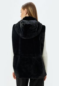 Schwarze Fake-Fur-Weste mit Kapuze, die eine weiche Textur und ein nahtloses Design aufweist. Der Rücken zeigt einen leicht längeren Schnitt und keine sichtbaren Verschlüsse.