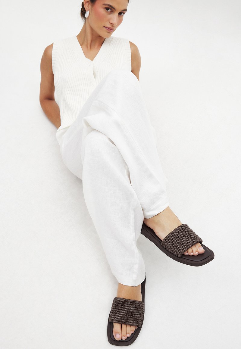 Donkerbruine slippers met een brede geweven band, platte zool, rechthoekige vorm en een gestructureerde afwerking, gecombineerd met witte linnen broek en een mouwloze top.