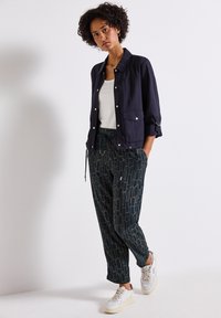 Una donna sta in piedi con sicurezza, indossando una giacca blu navy sopra una maglietta bianca e pantaloni a fantasia, completati da sneakers bianche su uno sfondo semplice.