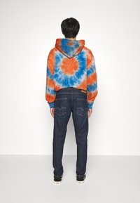 Sudadera de tie-dye en naranja y azul con un patrón circular; combinada con pantalones vaqueros de mezclilla oscuros con un corte clásico y bolsillos traseros visibles.