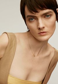 Top de cuero beige con tirantes anchos, que presenta un inserto de tela amarilla. Un delicado collar de oro con un pequeño colgante. Primer plano del escote.
