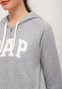 Sudadera gris con cremallera y logo blanco de "GAP", con capucha ajustable y bolsillos frontales. Tejido suave con una textura lisa.