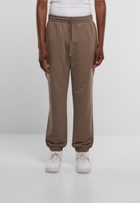Bruine joggers met elastische tailleband en gegroefde manchetten, ontworpen voor comfort, gecombineerd met witte sneakers op een effen achtergrond.