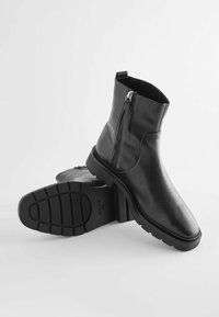 Bottines noires en cuir avec une texture lisse, dotées d'une fermeture éclair sur le côté, d'un bout arrondi et d'une semelle en caoutchouc épaisse et striée.