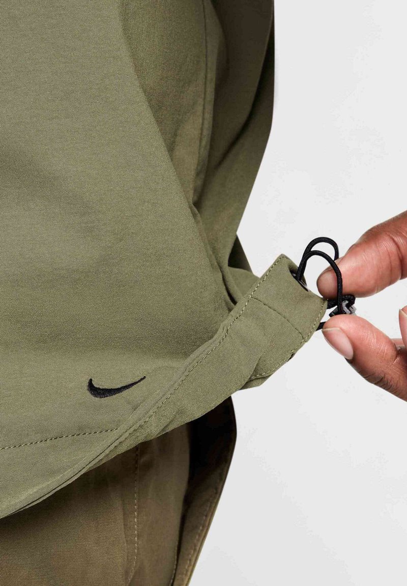 Olive zielona, lekka tkanina kurtki z czarnym logo Nike. Hem kurtki ma sznur do regulacji trzymany między palcami. Gładka faktura.