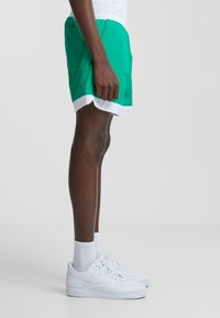 Grønne shorts med hvid kant, lavet af letvægtsmateriale, med sidelommer og elastisk linning. Sammen med hvide sneakers.