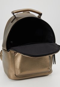 Metallisch bronzefarbener Rucksack in runder Form. Verfügt über einen Reißverschluss oben, ein schwarzes Innenfutter und eine Vordertasche mit glatter Textur.