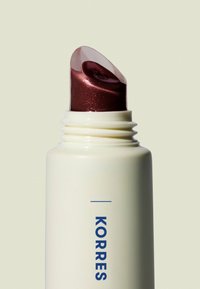 Producto labial de Korres en un tubo blanco con una marca azul; contiene una fórmula brillante, color burdeos, infundida con purpurina asomando por una tapa transparente.