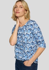 Blaue gemusterte Bluse mit runden Formen, weißen Akzenten und gerafften Ärmeln. Der Stoff hat eine glatte Textur und eine lockere Passform.