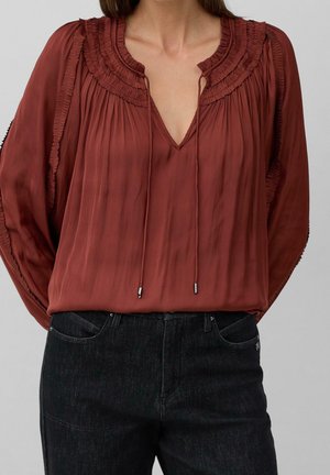 Vrouw draagt een roestkleurige blouse met lange mouwen met geplooide details en bindlinten, gecombineerd met een zwarte high-waisted spijkerbroek.