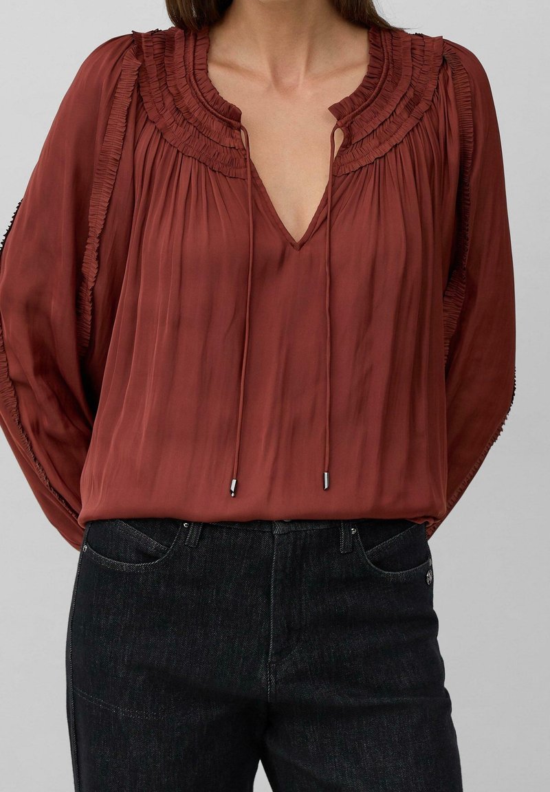 Femme portant un chemisier à manches longues couleur rouille avec des détails plissés et des liens à nouer, associé à un jean noir taille haute.