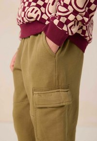 Le sweat-shirt présente des poignets côtelés bordeaux et un motif à carreaux crème amusant. Le pantalon vert olive a une poche latérale et une texture douce.