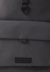 Sac à dos noir mat à l'avant avec fermeture à rabat et patch logo Tommy Hilfiger près du bas au centre.