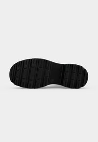Semelle de chaussure en caoutchouc noir avec des motifs de bande de roulement profonds et des sections de grip rectangulaires, montrée sur un fond gris clair uni.