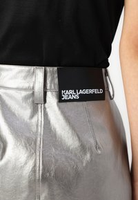 Jeans argintii metalici cu o textură netedă și o etichetă neagră pe care scrie "KARL LAGERFELD JEANS" cu litere albe îndrăznețe.
