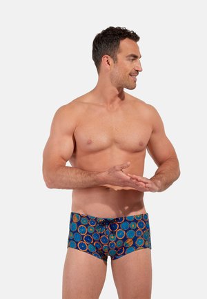 Oberkörperfreier Mann steht und lächelt seitlich, trägt blaue Badeshorts mit einem Muster aus orangefarbenen Zitrusfrüchten auf weißem Hintergrund.