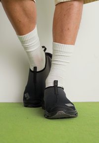 Fekete hálós slip-on cipők texturált felülettel, szürke talppal és fekete díszítésekkel, fehér bordás sportzoknival viselve.