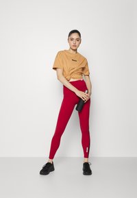 T-shirt de algodão laranja atado na cintura, combinado com leggings vermelhas de cintura alta. Sapatos de desporto pretos e uma garrafa de água escura na mão.
