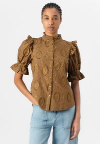 Blusa marrone ricamata con maniche a volant e collo alto; presenta intricate fantasie a foglia e chiusura con bottoni. Abbinata a jeans azzurri.
