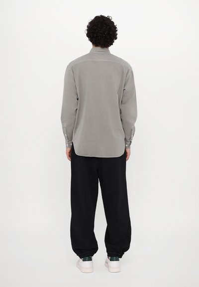 Chemise grise à manches longues avec un col boutonné, présentant une coupe droite et un ourlet arrondi, associée à un pantalon noir ample.