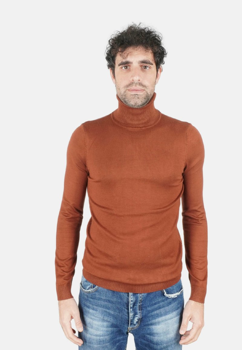 Maglione marrone aderente con collo e polsini a coste, indossato con jeans di jeans blu, su uno sfondo bianco semplice.