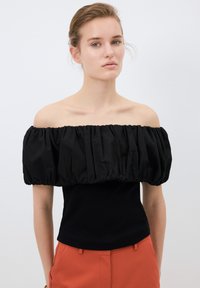 Top nero senza spalle con maniche a sbuffo arricciate e corpo a coste aderente. Texture liscia, design minimal, abbinato a pantaloni arancioni.