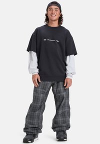 Felpa nera oversize con logo bianco, indossata sopra una maglietta grigia a maniche lunghe. Pantaloni baggy grigi a quadri, abbinati a scarpe nere.